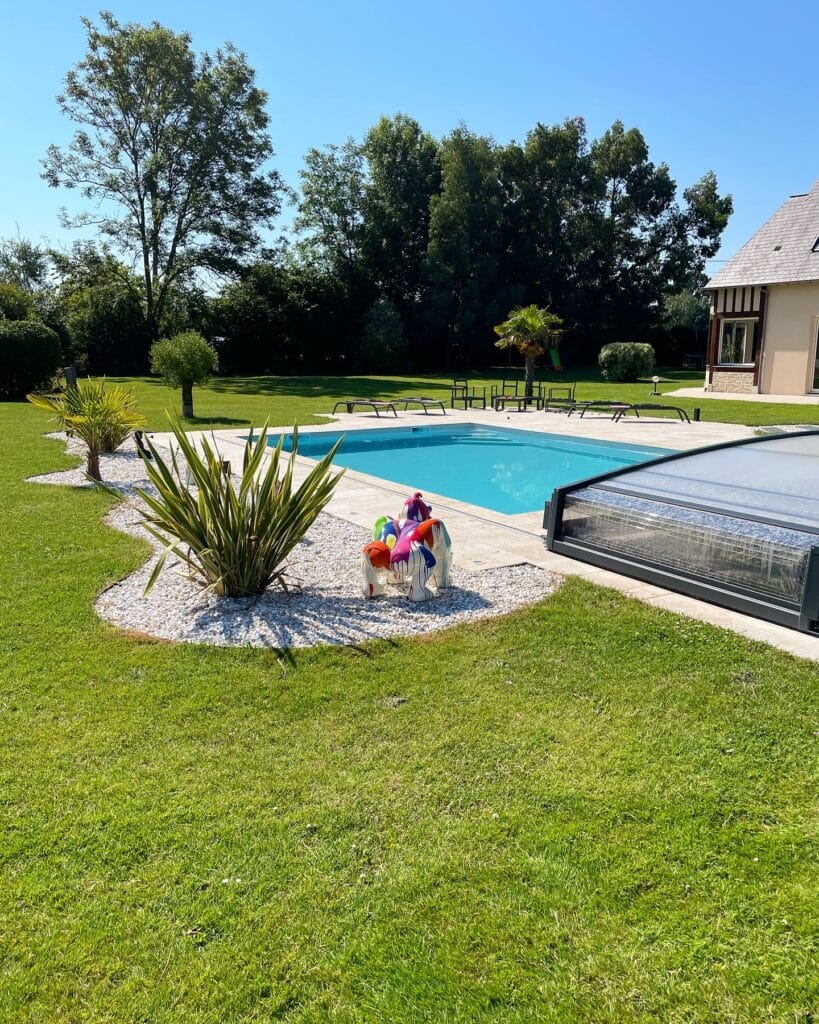 Piscine avec abri bas coulissant anthracite, massif de cordylines et galets blancs, chaises longues et maison normande en arrière-plan