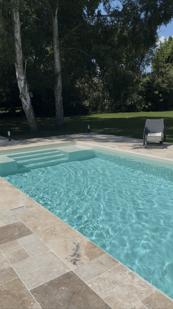 Piscine turquoise avec escalier intégré et terrasse en travertin beige dans un jardin arboré avec chaise longue