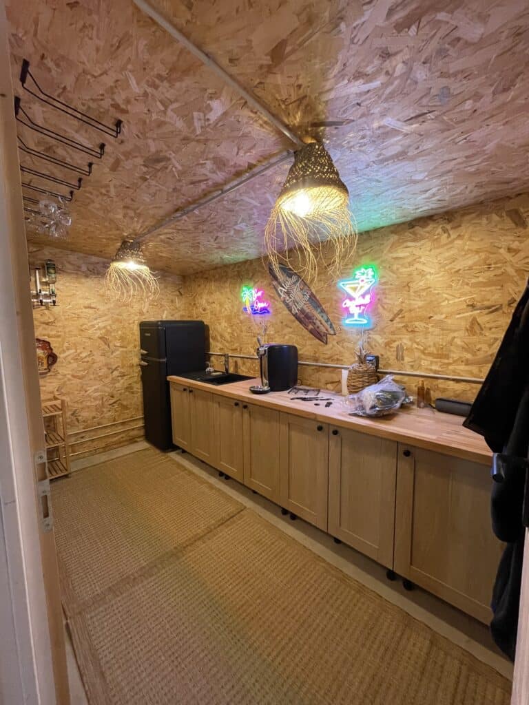 Intérieur de pool house aménagé en bar avec plan de travail bois, néons décoratifs, suspensions naturelles et murs en OSB