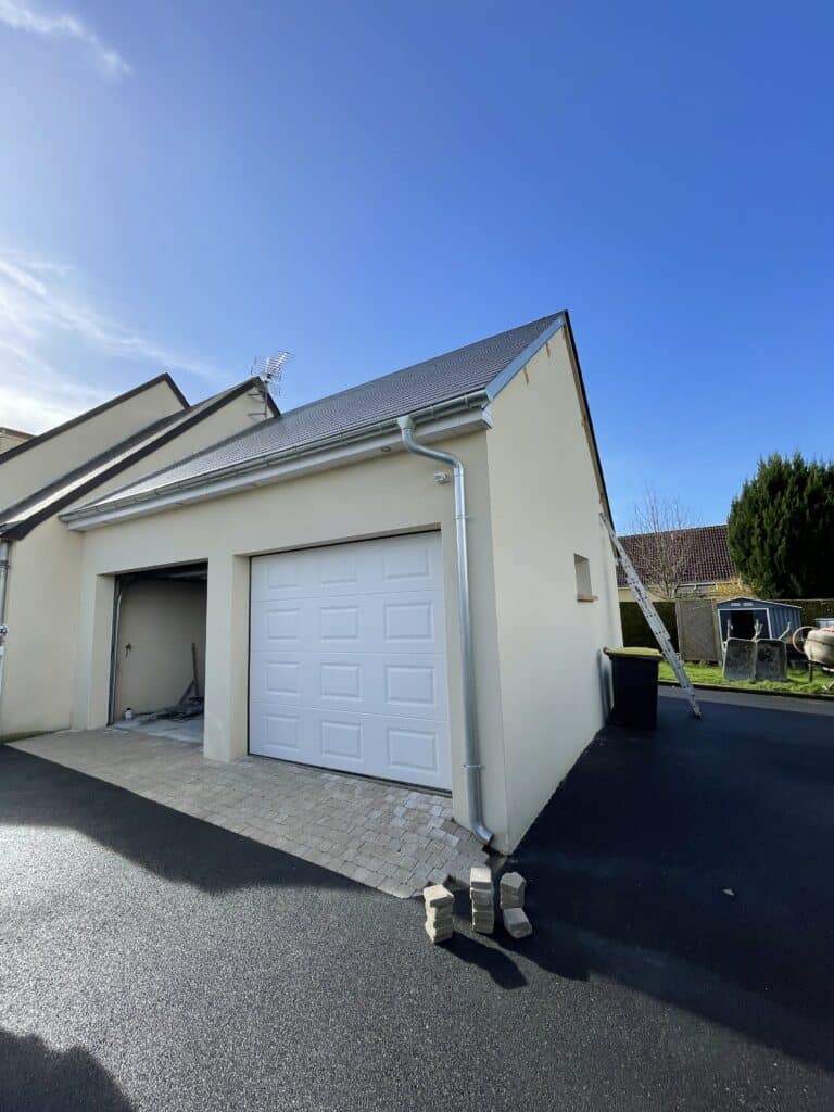 Extension garage avec porte sectionnelle blanche, toiture ardoise et façade enduite beige, allée en pavés et enrobé