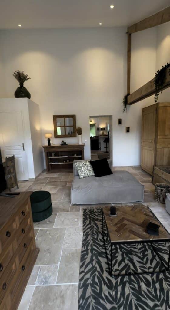 Salon rénové de grange avec sol en travertin, poutres apparentes, canapé gris et mobilier bois style campagne chic