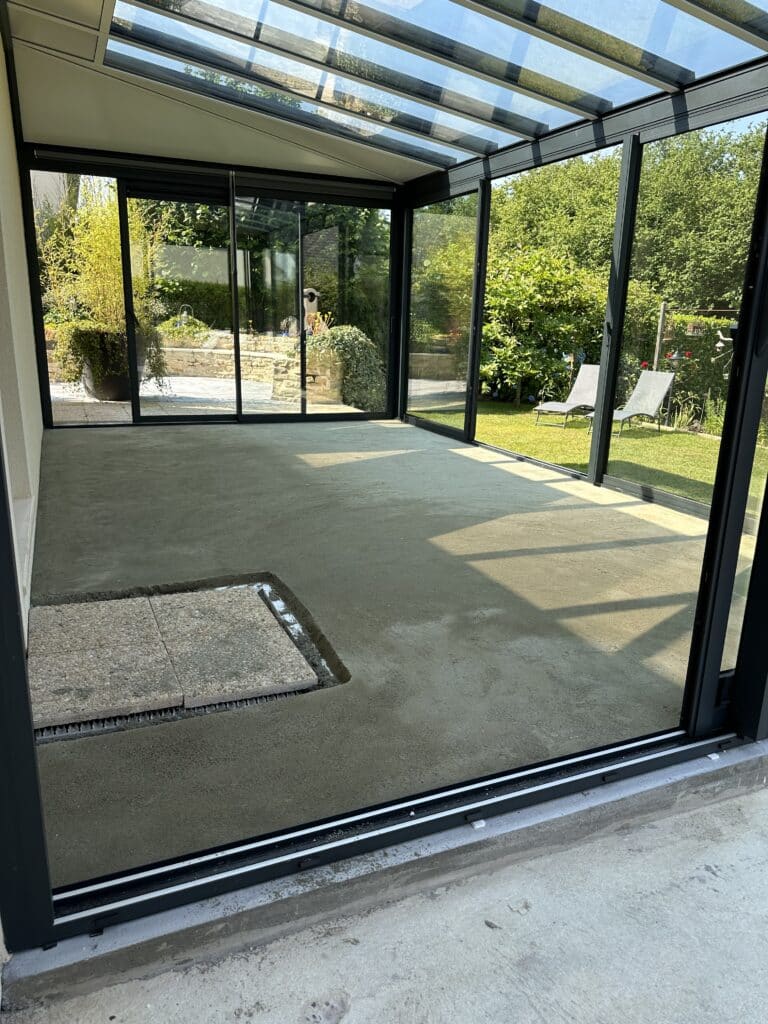 Intérieur de véranda en aluminium anthracite avec toit vitré en cours de dallage béton, vue sur jardin arboré et chaises longues