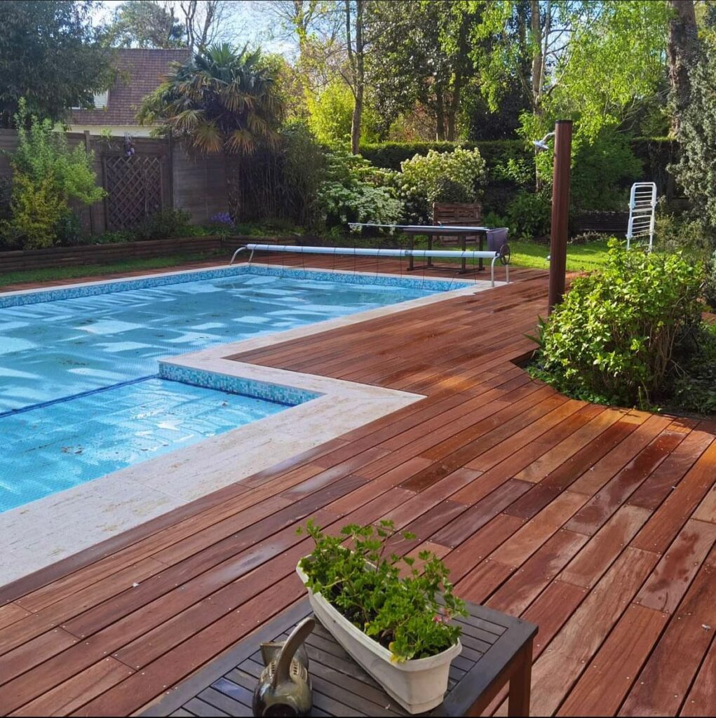 Piscine avec volet roulant et terrasse en bois exotique IPE rénové, entourée d'une végétation luxuriante au printemps