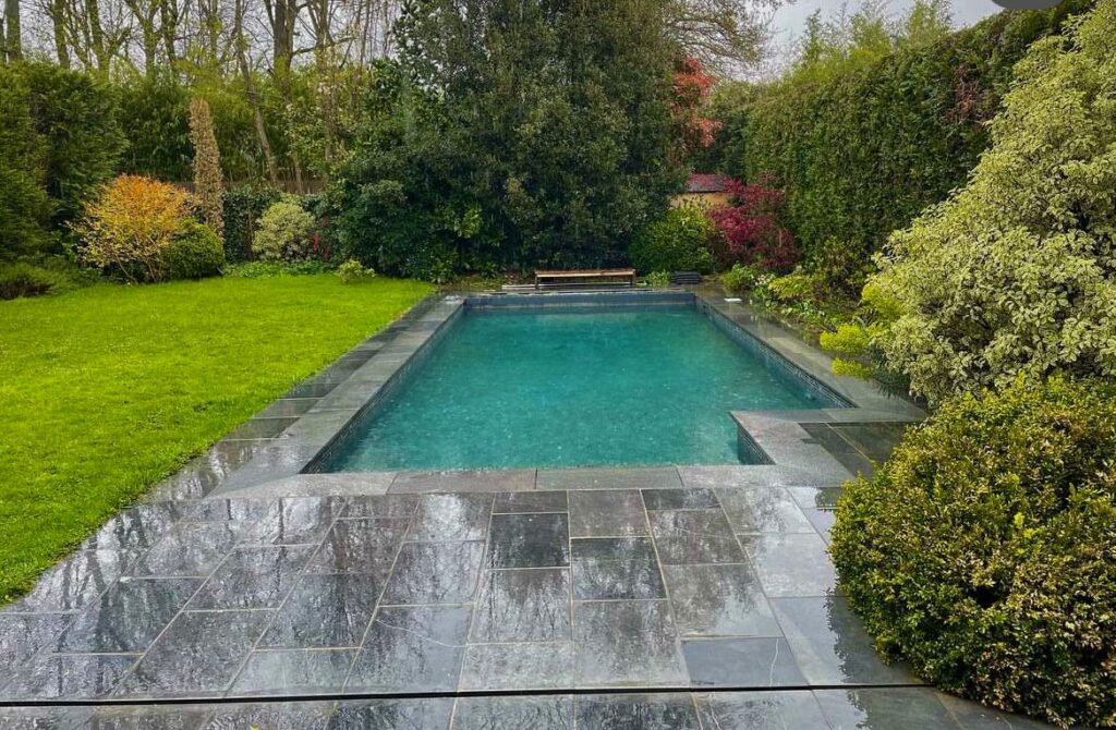 Piscine rectangulaire avec margelles en ardoise bleue et terrasse carrelée ardoise, intégrée dans un jardin arboré et fleuri