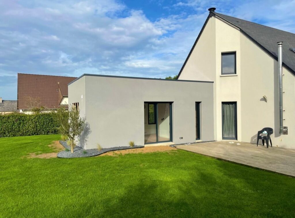 Extension moderne à toit plat en enduit gris avec baie vitrée, intégrée à une maison traditionnelle sur jardin pelousé