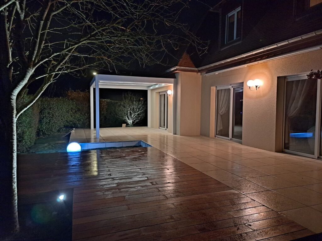 Terrasse en bois et carrelage avec piscine illuminée, pergola bioclimatique et boules lumineuses bleues de nuit