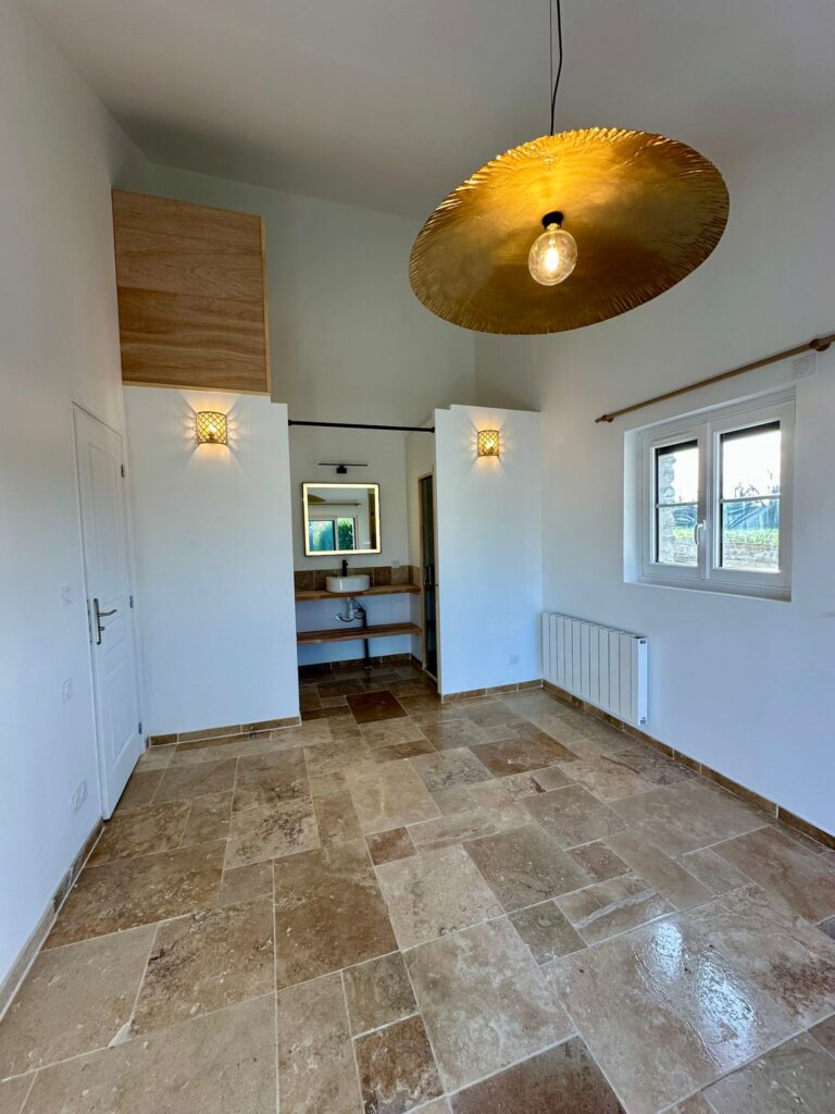 Chambre rénovée avec sol en travertin, suspension dorée design, appliques murales et salle de bain ouverte avec vasque à poser