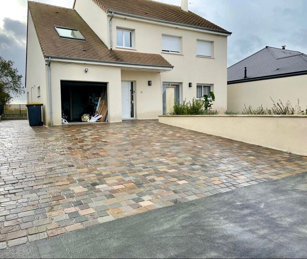 Allée et cour de maison pavée en pavés multicolores tons ocre et gris devant un pavillon avec garage ouvert