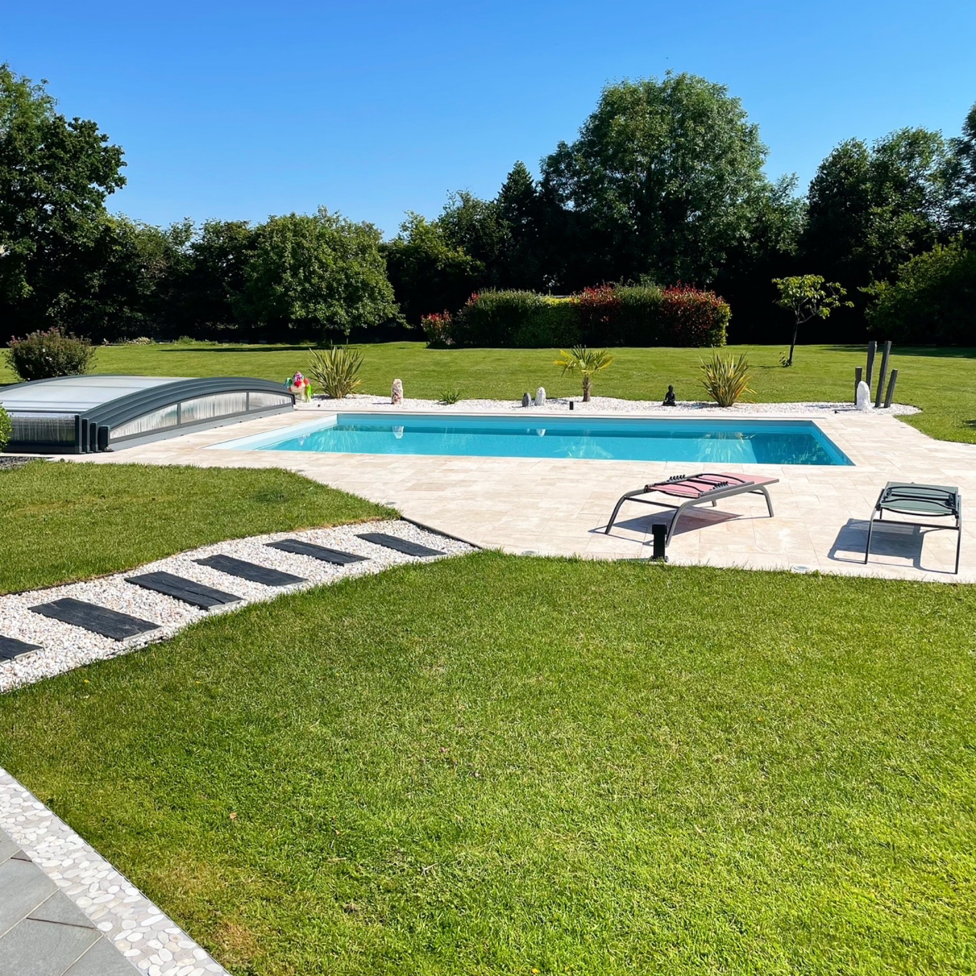 Piscine rectangulaire avec abri bas coulissant dans un jardin paysager avec pelouse et chaises longues